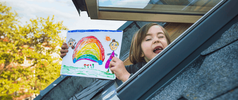 A boy sticks his head out the window with a rainbow drawing to symbolize COVID-19 optimism