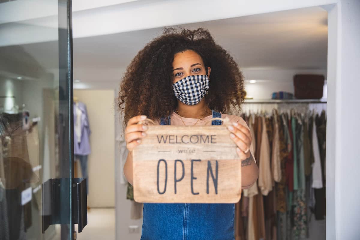 Woman with a mask, holding we're open sign in a store doorway