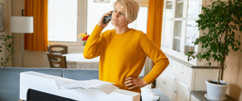 A frustrated woman on the phone with a new television in a box in front of her. Trying to return a product.