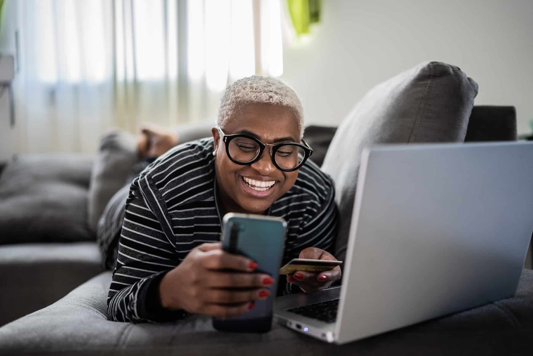 A smiling woman shopping online while laying on a couch