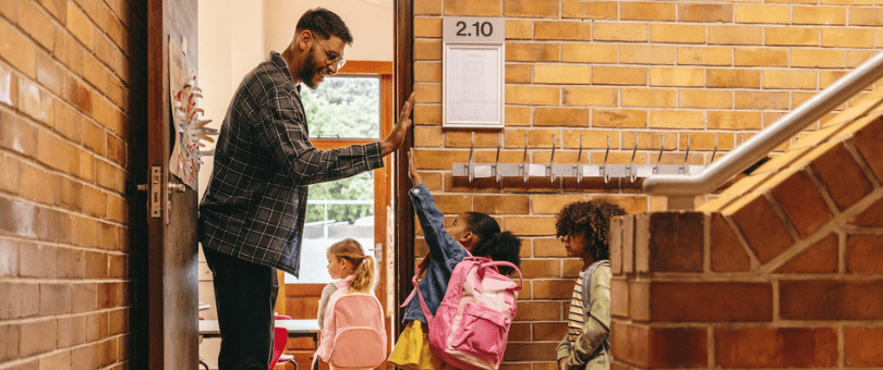 A tall teacher welcoming his very young students back to school with high fives.