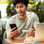 man shopping on his laptop at a cafe, checking his phone perhaps for an OTP to illustrate Signifyd 3D Secure explainer