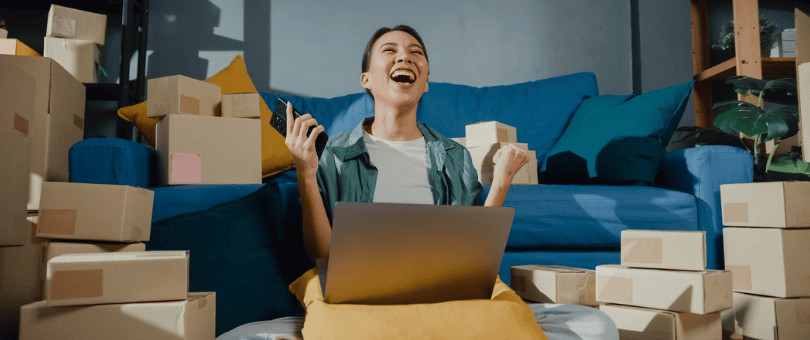 A woman gleeful at her laptop amid piles of boxes from ecommerce orders — to illustrate malicious friendly fraud and gaps in AI fraud detection.